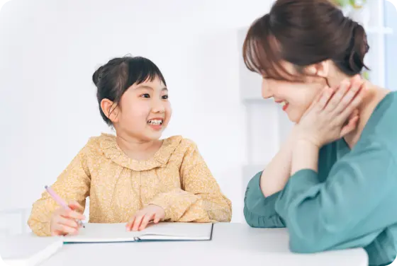 テーブルに座る女性と小さな女の子が、楽しそうに一緒に書き物をしているシーン。